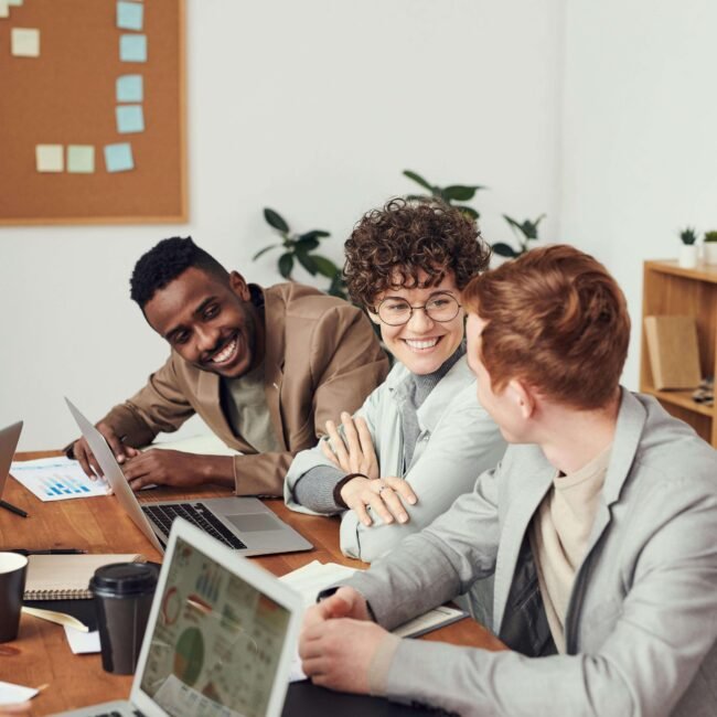 Groupe de collègues souriants travaillant ensemble autour d’une table avec ordinateurs et graphiques, illustrant le travail d’équipe dans une stratégie d’entreprise